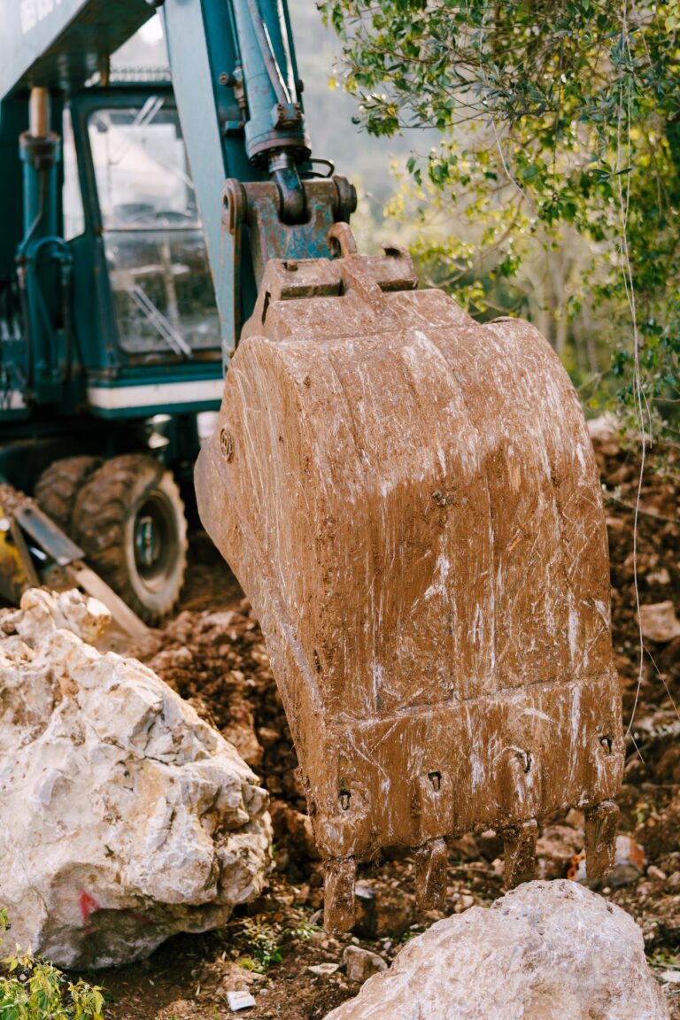 Close-up of a muddy excavator bucket at a construction site, surrounded by rocks and earth.